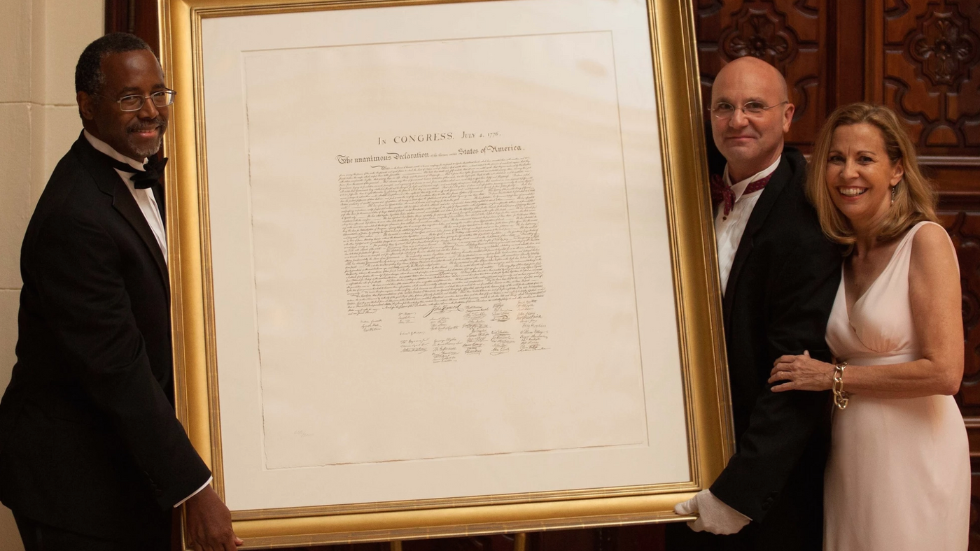 Our Founding Document Dr. Ben Carson with Paul and Sara Fattori holding up a Federal Style gold gilded frame around the hand engraved facsimile of the unanimous declaration of independence by artist jose maria cundin